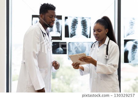 Male and female doctors discussing patient diagnosis, consulting x-ray images at clinic window. Collaborative healthcare professionals analyzing medical information to provide accurate treatment plan. Male and female doctors discussing patient diagnosis, consulting x-ray images at clinic window. Collaborative healthcare professionals analyzing medical information to provide accurate treatment plan. 130329257
