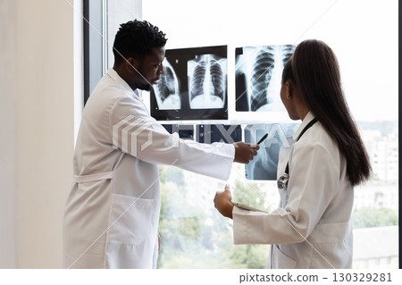 Male and female doctors of diverse backgrounds examining chest X-ray images by window. Collaborative medical consultation in modern clinic setting, emphasizing teamwork, analysis Male and female doctors of diverse backgrounds examining chest X-ray images by window. Collaborative medical consultation in modern clinic setting, emphasizing teamwork, analysis 130329281