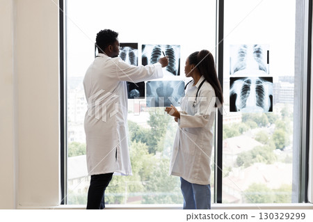 Medical professionals, male and female, reviewing x-ray diagnoses near a bright clinic window. Collaborative discussion over radiological findings indicates teamwork for accurate analysis. Medical professionals, male and female, reviewing x-ray diagnoses near a bright clinic window. Collaborative discussion over radiological findings indicates teamwork for accurate analysis. 130329299