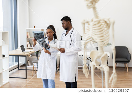 Focused male and female doctors analyzing X-ray images, medical skeleton in foreground, study environment. Focused male and female doctors analyzing X-ray images, medical skeleton in foreground, study environment. 130329307