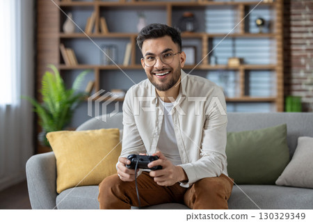 Hispanic man sitting on comfortable sofa holding gaming controller, engaging in leisure activity. Casual setting reflects enjoyment, relaxation, and entertainment at home. 130329349