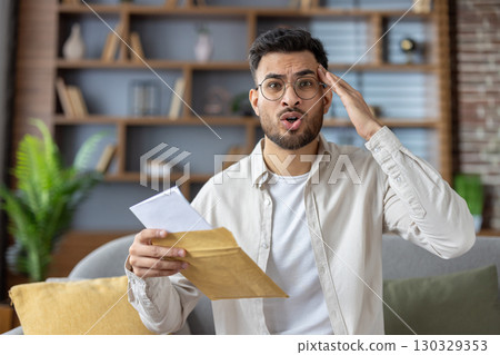 Surprised man with glasses holding envelope showing shock. He appears to receive unexpected news while sitting on couch in living room. Casual attire reflects relaxed and homey atmosphere 130329353