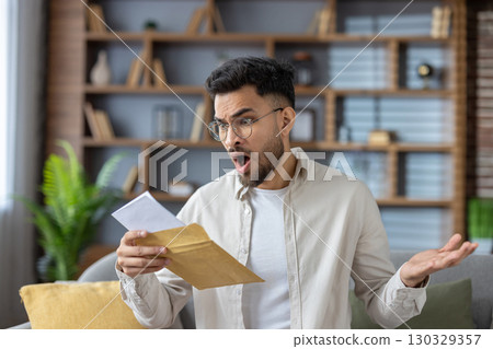 Hispanic man experiencing surprise while opening letter at home. Captured indoors with expressive face, conveying emotions related to unexpected news or surprise. Suitable for concepts of 130329357