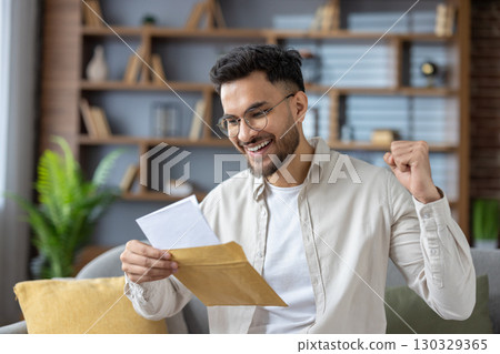 Close-up photo of a young indian man in a shirt and bib sitting on the couch at home and reading a letter he received, happy with the news and message. 130329365