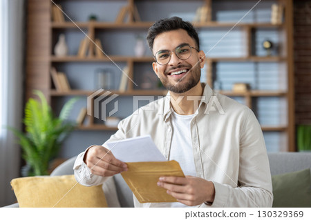 Smiling man opening letter while sitting on sofa in modern living room. Radiating happiness and positive emotions, creating warm and welcoming atmosphere. Casual and relaxed indoor setting. Smiling man opening letter while sitting on sofa in modern living room. Radiating happiness and positive emotions, creating warm and welcoming atmosphere. Casual and relaxed indoor setting. 130329369
