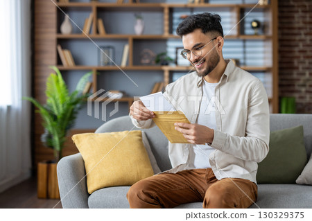 Hispanic man sitting on sofa enjoying pleasant surprise from envelope. Relaxed and happy, surrounded by home decor. Concept of positive communication and personal moments. Hispanic man sitting on sofa enjoying pleasant surprise from envelope. Relaxed and happy, surrounded by home decor. Concept of positive communication and personal moments. 130329375