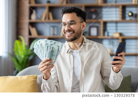 Smiling man with glasses holding cash in one hand and smartphone in other. Shows happiness and financial success in comfortable living room setting. 130329378