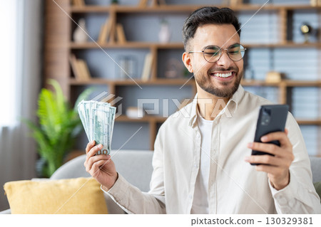 Smiling man holding dollar bills, using smartphone, expressing success and financial achievement. Depicts modern lifestyle, digital technology, financial gain. Smiling man holding dollar bills, using smartphone, expressing success and financial achievement. Depicts modern lifestyle, digital technology, financial gain. 130329381