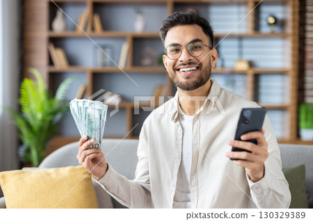 Confident man with glasses sitting on sofa holding cash and smartphone, symbolizing success in finance or online business. Happy and motivated expression highlights achievement and prosperity 130329389