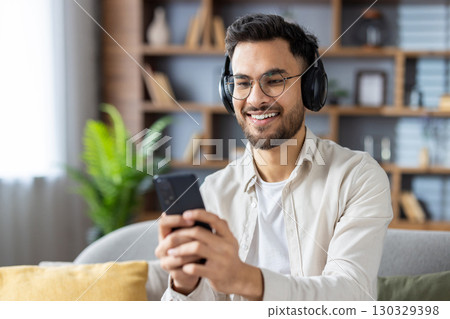 Smiling man wearing headphones using smartphone, possibly enjoying music or chatting online. Seated on couch in cozy home environment, expressing relaxation and enjoyment. Smiling man wearing headphones using smartphone, possibly enjoying music or chatting online. Seated on couch in cozy home environment, expressing relaxation and enjoyment. 130329398