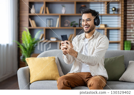 Man wearing headphones sits on sofa using smartphone, enjoying music or video. Captures mood of relaxation, modern technology, digital lifestyle, and immersive audio experience. Man wearing headphones sits on sofa using smartphone, enjoying music or video. Captures mood of relaxation, modern technology, digital lifestyle, and immersive audio experience. 130329402