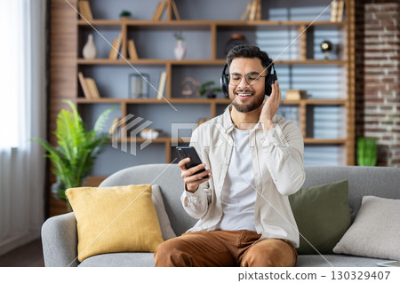 Smiling and relaxed young Indian man sitting at home on the sofa wearing headphones, holding the phone in his hands, resting with his eyes closed. Smiling and relaxed young Indian man sitting at home on the sofa wearing headphones, holding the phone in his hands, resting with his eyes closed. 130329407