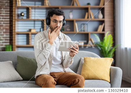 Young man sitting on sofa using tablet and headphones, looking surprised. Modern living room. Comfortable and leisure setup, with plants in background, showcasing technological engagement. 130329410