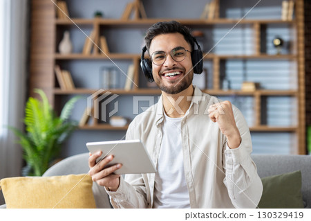 Confident man with headphones and tablet smiling and clenching fist in excitement. Seated on sofa, he celebrates success or positive news while enjoying music or an online interaction with tablet. 130329419
