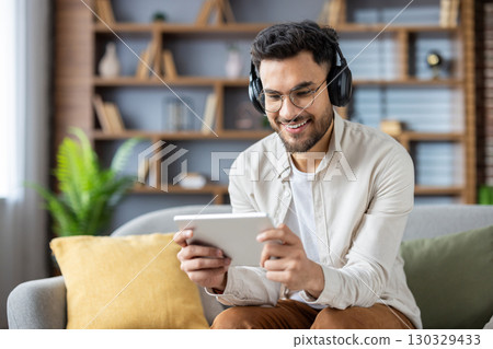 Cheerful man wearing headphones and casual clothes sits on sofa using digital tablet for entertainment or education. Relaxed, happy atmosphere with modern home setting enhances tech-focused lifestyle 130329433