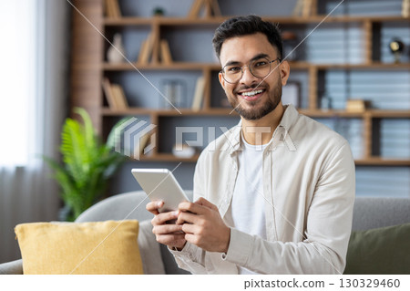 Cheerful man with glasses holding tablet sitting on sofa enjoying digital content. Modern living room setting conveys relaxation and home comfort. Young male engages with technology, showcasing Cheerful man with glasses holding tablet sitting on sofa enjoying digital content. Modern living room setting conveys relaxation and home comfort. Young male engages with technology, showcasing 130329460