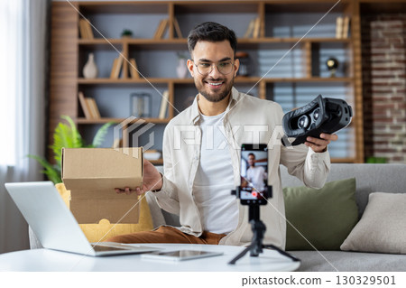 Young man creating unboxing video using smartphone. He holds virtual reality headset and cardboard box, capturing engaging content in home setting. Enthusiastic expression shows excitement about tech Young man creating unboxing video using smartphone. He holds virtual reality headset and cardboard box, capturing engaging content in home setting. Enthusiastic expression shows excitement about tech 130329501