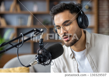 Young man wearing headphones speaking into microphone in podcast studio. He appears focused and engaged in serious conversation. Modern equipment and professional setup suggest tech-savvy environment 130329507