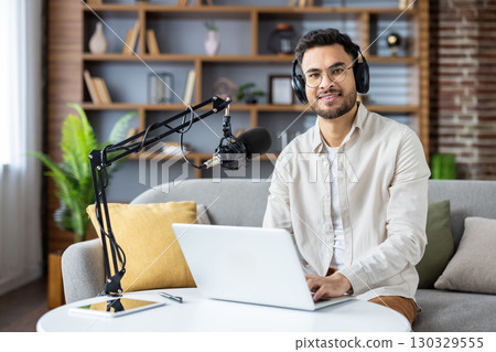 Smiling podcaster using laptop and microphone in home studio. Headphones on, sitting at desk, creating audio content with enthusiasm. Modern setup enhances recording experience, Smiling podcaster using laptop and microphone in home studio. Headphones on, sitting at desk, creating audio content with enthusiasm. Modern setup enhances recording experience, 130329555