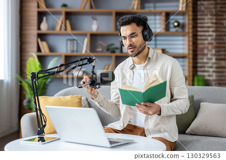 Hispanic man engaged in podcast recording at home using microphone, laptop, and headphones. Sitting on couch, holding book, and speaking enthusiastically. Ideal for concepts like podcasting 130329563