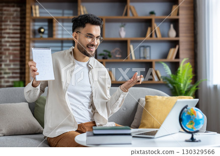 Cheerful online teacher conducting virtual class from home. Man with glasses gesturing while holding notebook, engaging with students through laptop. 130329566