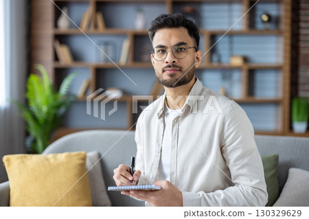 Portrait of a young Indian man sitting on the sofa at home, holding a notebook and a pen in his hands, looking seriously and confidently at the camera. Portrait of a young Indian man sitting on the sofa at home, holding a notebook and a pen in his hands, looking seriously and confidently at the camera. 130329629