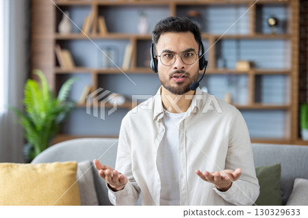 Portrait of a young Indian man wearing glasses and a headset sitting on the couch at home and talking to the camera, while gesturing with his hands. Portrait of a young Indian man wearing glasses and a headset sitting on the couch at home and talking to the camera, while gesturing with his hands. 130329633