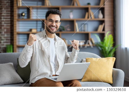 Portrait of a happy Indian man rejoicing in success and good news, sitting on the couch at home, making a winning gesture with his hands and looking at the camera with a smile. 130329638