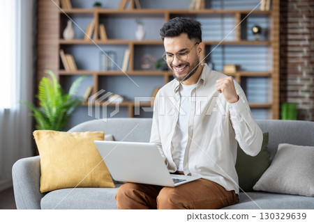 Happy young Indian man sitting on sofa at home, using laptop on lap, looking smiling at screen, showing victory yes with hand. Happy young Indian man sitting on sofa at home, using laptop on lap, looking smiling at screen, showing victory yes with hand. 130329639