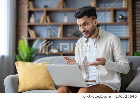 Serious young Indian nightingale sitting on sofa at home, holding laptop on lap and talking remotely on video call, gesturing with hands. Serious young Indian nightingale sitting on sofa at home, holding laptop on lap and talking remotely on video call, gesturing with hands. 130329640