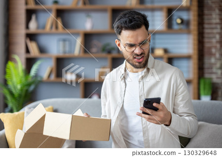 Angry young Indian man sitting at home holding mistakenly received package and looking worriedly at mobile phone screen. 130329648