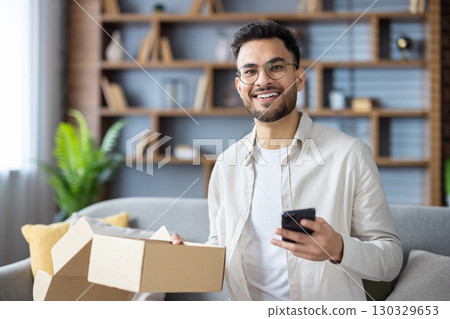 Portrait of a happy young Indian man sitting on the couch at home, holding a received parcel and a mobile phone, smiling and looking at the camera. 130329653