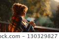 A young Caucasian woman with curly hair sits on a wooden porch wrapped in a plaid blanket, holding a steaming cup, enjoying the autumn sunlight. 130329827