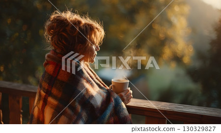 A young Caucasian woman with curly hair sits on a wooden porch wrapped in a plaid blanket, holding a steaming cup, enjoying the autumn sunlight. 130329827