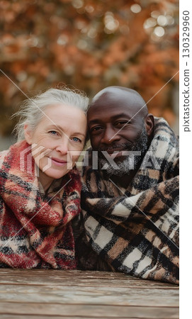 An interracial couple enjoys a cozy moment in autumn. The woman has long gray hair and fair skin. The man has a bald head and a beard, with dark skin. They are wrapped in blankets. 130329960