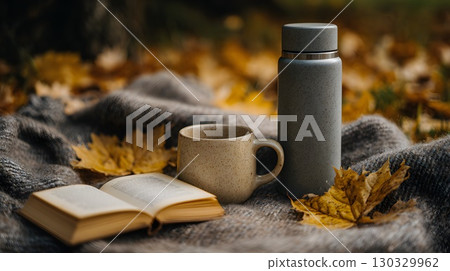 A cozy autumn picnic scene with a thermos, a cup, and an open book on a blanket surrounded by fallen leaves in a park. A cozy autumn picnic scene with a thermos, a cup, and an open book on a blanket surrounded by fallen leaves in a park. 130329962