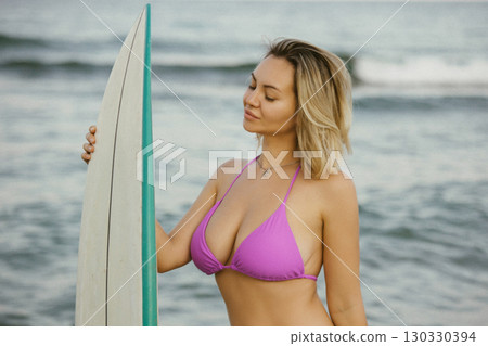Attractive young woman in a purple bikini posing with a surfboard on the beach, enjoying summer by the sea, confident and relaxed, ocean waves in the background. Attractive young woman in a purple bikini posing with a surfboard on the beach, enjoying summer by the sea, confident and relaxed, ocean waves in the background. 130330394