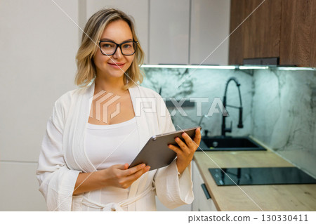 Confident woman in glasses and bathrobe holding tablet in modern kitchen. Real estate agent or interior designer working from home, smart casual look, stylish interior in background. Confident woman in glasses and bathrobe holding tablet in modern kitchen. Real estate agent or interior designer working from home, smart casual look, stylish interior in background. 130330411