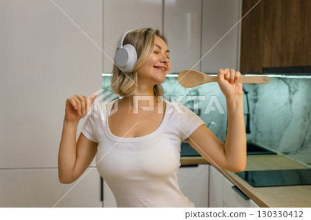 Happy young woman singing with wooden spoon like microphone, wearing headphones in modern kitchen. Joyful moment, music and fun at home. Happy young woman singing with wooden spoon like microphone, wearing headphones in modern kitchen. Joyful moment, music and fun at home. 130330412