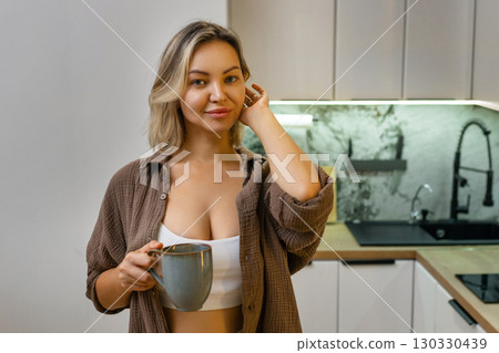 Modern kitchen: Young woman in a white crop top and open shirt stands with a mug in hand, enjoying a peaceful morning at home, relaxed and smiling. Modern kitchen: Young woman in a white crop top and open shirt stands with a mug in hand, enjoying a peaceful morning at home, relaxed and smiling. 130330439