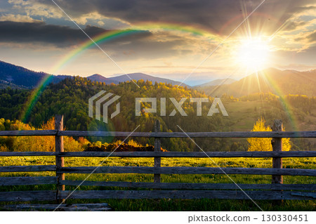 trees on the hill and fence on the grassy field at sunset. spring rural landscape in evening light. distant mountain ridge beneath sky with clouds. idyllic alpine farmland under the rainbow trees on the hill and fence on the grassy field at sunset. spring rural landscape in evening light. distant mountain ridge beneath sky with clouds. idyllic alpine farmland under the rainbow 130330451