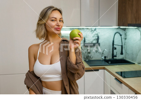 Attractive young woman in crop top playfully poses with a green apple in modern kitchen. Healthy lifestyle, flirtatious mood, natural beauty, wellness and nutrition vibes. 130330456