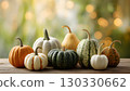 A collection of various pumpkins in different colors and sizes arranged on a wooden table. The background features soft, blurred lights, creating a festive atmosphere. 130330662