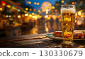 A glass of beer sits on a wooden table with sausages. The background features a festive atmosphere with lights and a ferris wheel, typical of Oktoberfest in Bavaria, Germany. 130330679