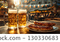 Two large beer mugs filled with golden beer sit on a wooden table next to a plate of grilled sausages. The background features festive decorations typical of Oktoberfest. 130330683