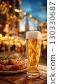 A glass of golden beer with a frothy head sits on a wooden table next to a plate of sausages. The background features festive lights and a blurred cityscape. 130330687