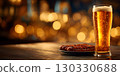 A tall glass of golden beer sits on a wooden table next to a plate of pretzels. The background features warm, blurred lights, evoking a festive atmosphere. 130330688