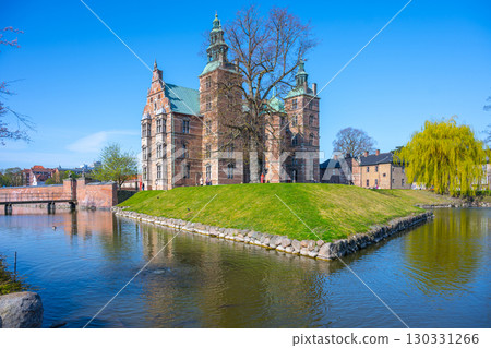 Rosenborg Castle showcases its stunning renaissance architecture in Copenhagen, surrounded by tranquil waters and lush greenery on a bright, clear day with blue skies. 130331266