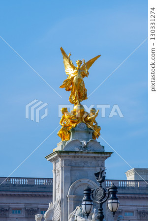 The elegant Queen Victoria Memorial stands proudly in front of Buckingham Palace, featuring a majestic golden angel surrounded by sculptures against a clear blue sky. 130331274