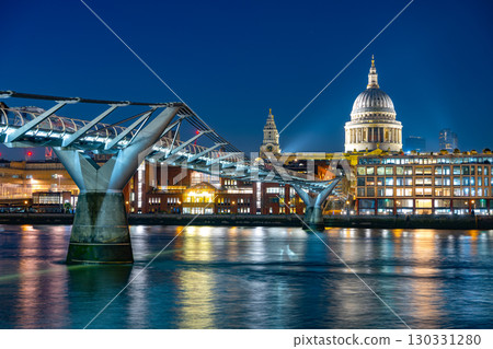 As evening falls, the Millenium Bridge elegantly spans the River Thames, showcasing stunning reflections while St. Paul's Cathedral stands majestically illuminated in the background. 130331280